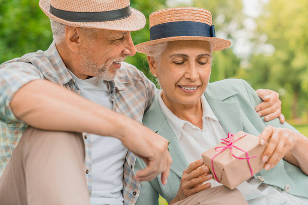 Cheerful aged man looking at his wife while giving a nice present box to her for a birthday - Фото, изображение