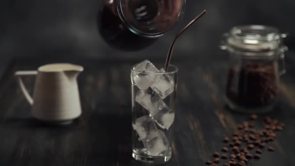 Add black coffee of cream being poured into a glass of cold brew iced coffee on black wood table with coffee beans on black background - Footage, Video