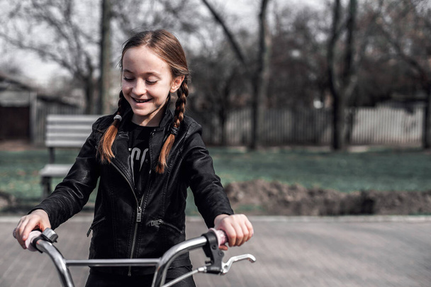 Meisje op een fietstocht. Portret van een meisje met vlechten in een leren jasje op een fiets. Het meisje is blij en glimlacht. Sportactiviteiten - Foto, afbeelding