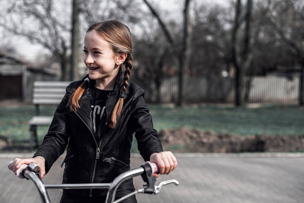 Girl on a bike ride. Portrait of a girl with braids in a leather jacket on a bicycle. The girl is joyful and smiling. Sports activity - Zdjęcie, obraz