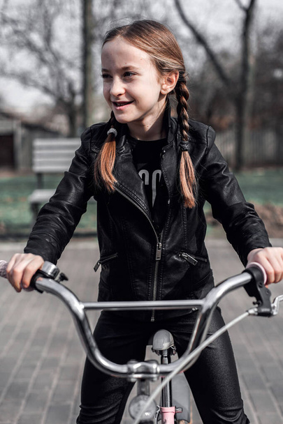 Una chica en un paseo en bicicleta. Retrato de una chica con trenzas en una chaqueta de cuero en una bicicleta. La chica es alegre y sonriente. Actividad deportiva
 - Foto, Imagen