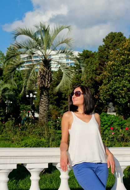 Nice woman walk at city enjoys vacation. Flowers garden with palm at background. Cheerful, smiling, lucky lady in white blouse, blue trousers and sunglasses outdoors. Batumi, Georgia. - 写真・画像