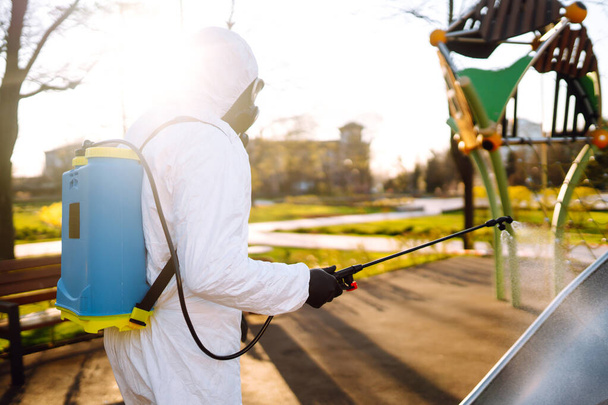 Man wearing protective suit disinfecting the playground in the sun with spray chemicals to preventing the spread of coronavirus, pandemic in quarantine city. Covid -19. Cleaning concept. - 写真・画像
