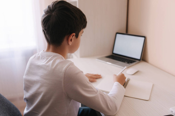 Iskolásfiú tanul az íróasztalnál a szobájában. A srác laptopot használ és jegyzetel. Könyvek és táblagép az asztalon. Tanulj otthon a qurantine alatt - Fotó, kép