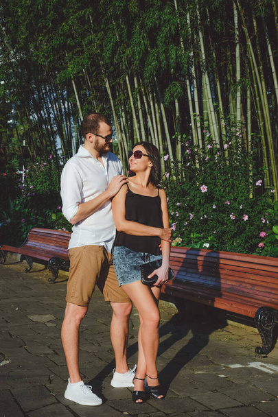 Happy couple of lovers walking outdoors. Bamboo gardens at background. Cheerful, smiling man and lucky lady in sunglasses have fun time in summer. - Фото, зображення
