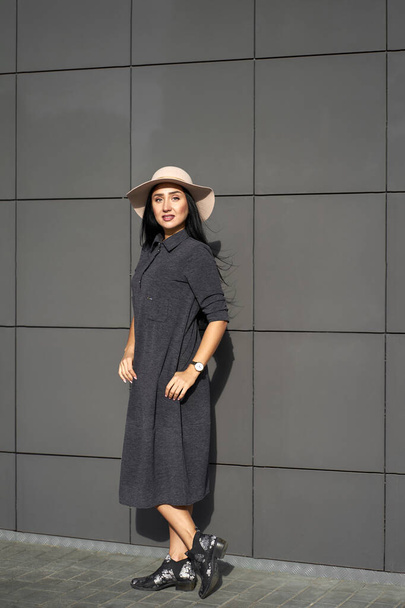 Aantrekkelijk modemodel met stijlvolle grijze jurk en modieuze hoed poserend op grijze muurachtergrond. Outdoor portret.                               - Foto, afbeelding