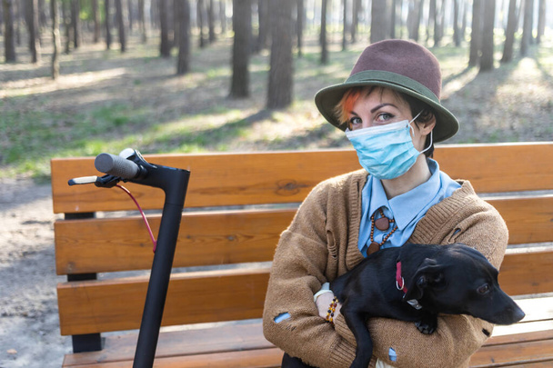 Una mujer elegante con máscara y guantes para defenderla de coronavirus. Usar scooter eléctrico en la ciudad y aparcar para estar a salvo. Divirtiéndose con su perro
. - Foto, imagen