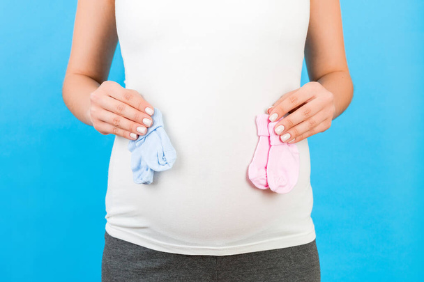 Oříznutý portrét těhotné ženy v domácím oblečení drží dítě růžové a modré ponožky na břiše na modrém pozadí. Je to kluk nebo holka, co čeká porod. Kopírovat prostor. - Fotografie, Obrázek