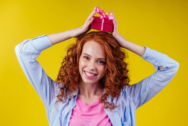 redhair curly ginger woman holding gift box on her head in studio yellow background.mothers day and eighth of marchor St. Valentines Day concept - Photo, Image