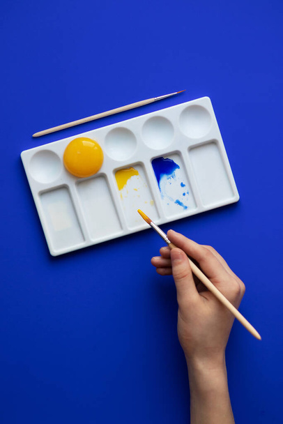 White palette with watercolor paints. Hands hold brushes. Blue background. Instead of yellow, egg yolk is used. Creative design and art. Top view and copy space. - Foto, afbeelding