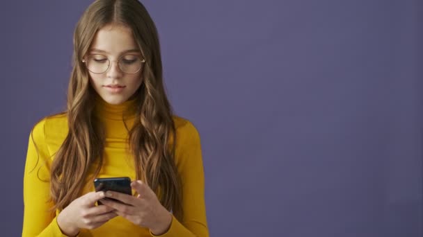 Spokojna, miła nastolatka używa swojego smartfona stojącego na fioletowym tle w studio - Materiał filmowy, wideo