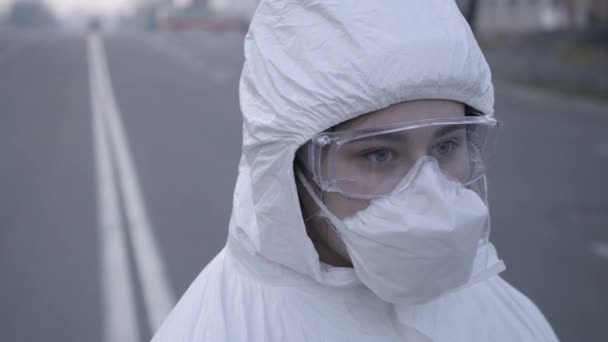 Close-up of sad Caucasian woman in protective antiviral suit turning eyes to camera. Portrait of young doctor or virologist controlling coronavirus quarantine on empty city road. Covid-19 pandemic. - Video, Çekim