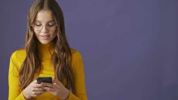 A pleased smiling teenage girl is using her smartphone standing isolated over a purple background in studio - Materiał filmowy, wideo