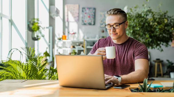 Front view shot of smiling young businessman drinking coffee while using laptop in office - Foto, immagini