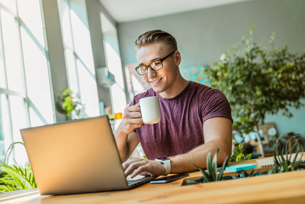 Веселый молодой бизнесмен пьет кофе во время использования ноутбука в офисе
 - Фото, изображение