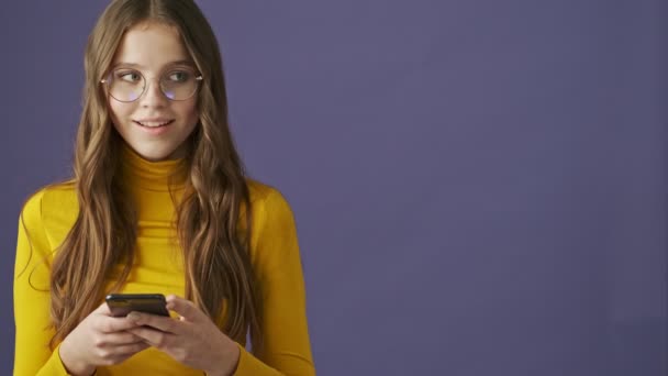 A charming satisfied teenage girl is using her smartphone standing isolated over a purple background in studio - Footage, Video