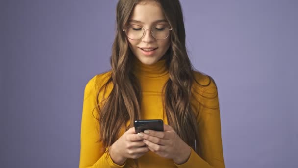 A positive smiling teenage girl is using her smartphone standing isolated over a purple background in studio - Felvétel, videó