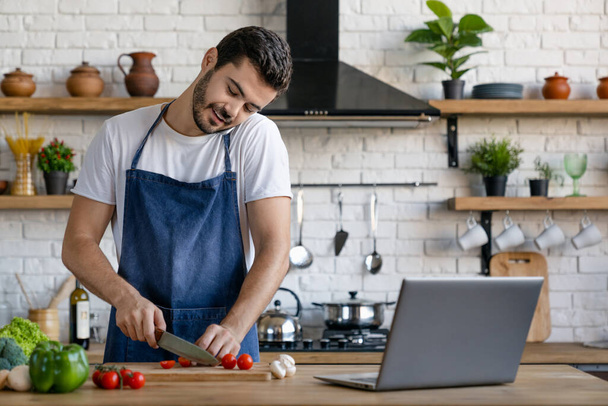 Красивый улыбающийся кавказский мужчина готовит на кухне, разговаривая по мобильному телефону с ноутбуком на столе
 - Фото, изображение