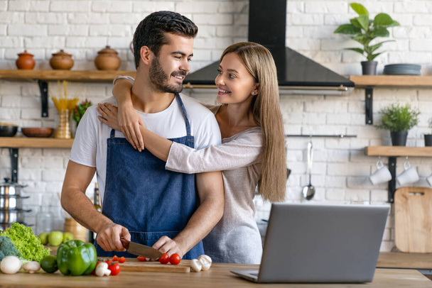 Beautiful cheerful young couple cooking dinner with laptop on table while standing in the kitchen at home - Фото, изображение