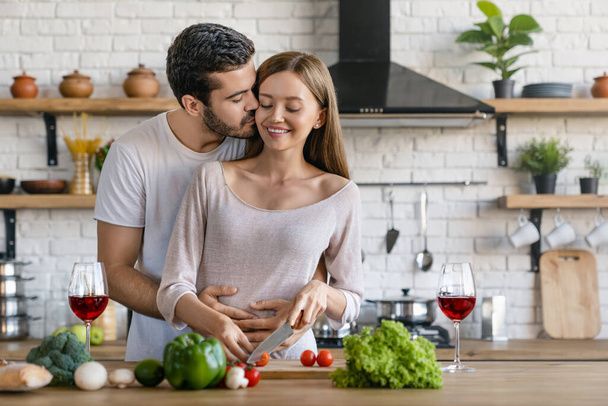 Beautiful cheerful young couple cooking dinner while man kissing his girlfriend standing in the kitchen at home - Foto, immagini