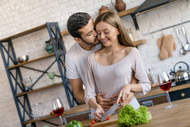 Beautiful cheerful young couple cooking dinner while man kissing his wife standing in the kitchen at home - Foto, Bild