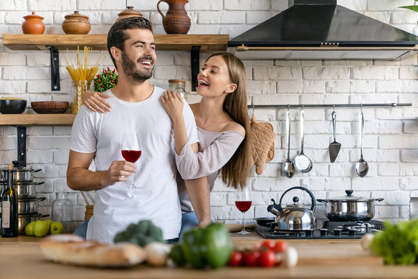 Beautiful laughing young couple drinking wine and cooking salad while standing in the kitchen - Valokuva, kuva