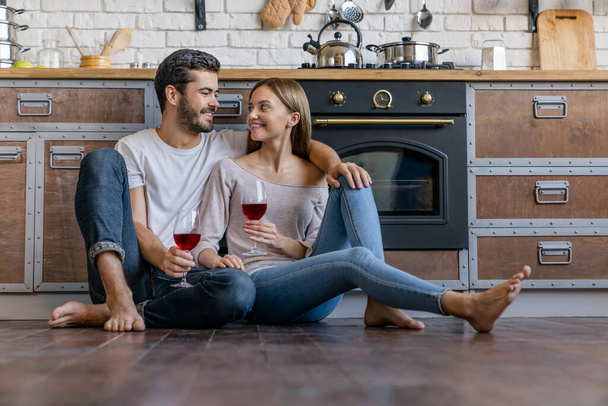 Happy young man and woman sitting on kitchen floor holding wine glasses - Fotografie, Obrázek