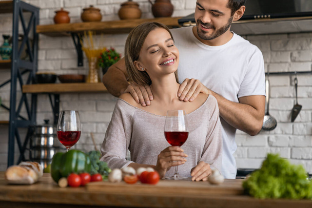 Beautiful girl drinking wine while handsome man making massage to her in home kitchen - Foto, Imagen
