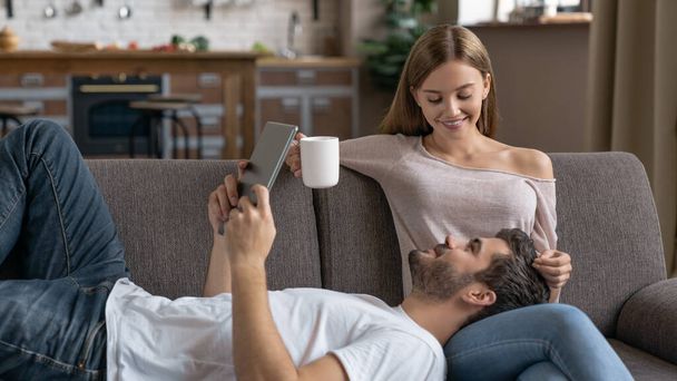 Joven pareja feliz tumbada en el sofá con tableta digital y taza de café
 - Foto, imagen