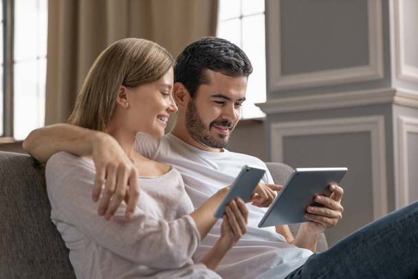 Side view of young happy couple hugging and using smartphone, digital tablet while sitting on sofa at home - Фото, зображення