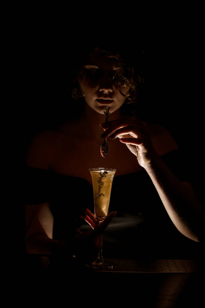 young attractive barman girl at dark bar holds spoon over glass with cocktail - Фото, изображение