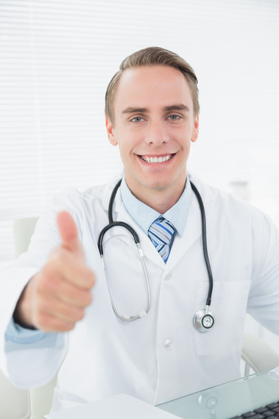Smiling doctor gesturing thumbs up at medical office - Fotó, kép