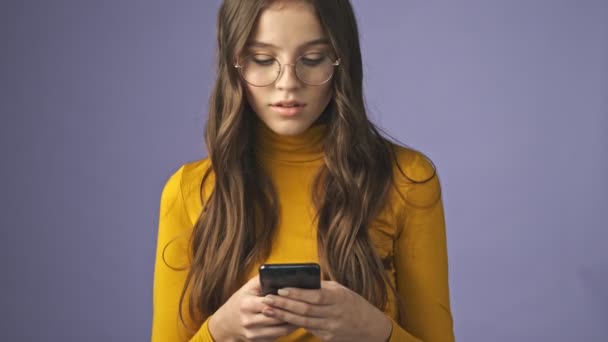 zaneprázdněný dospívající dívka je chatování na její smartphone stojící izolované přes fialové pozadí ve studiu - Záběry, video