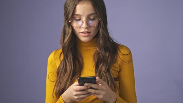 A busy teenage girl is chatting on her smartphone while recognize someone and waving hand to him standing isolated over a purple background in studio - Footage, Video