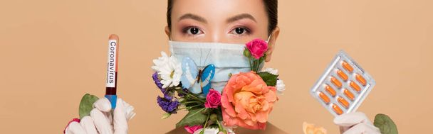 панорамный снимок азиатской девушки в латексных перчатках и цветочной маске для лица с бабочкой, держащей таблетки и коронавирусный анализ крови, выделенный на бежевом
 - Фото, изображение
