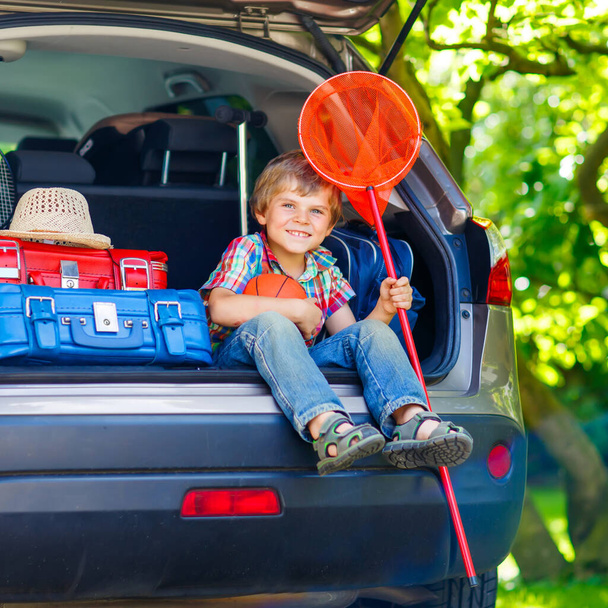 Ενθουσιασμένο αγοράκι που κάθεται στο πορτ-μπαγκάζ του αυτοκινήτου λίγο πριν φύγει για καλοκαιρινές διακοπές με την οικογένειά του. Ευτυχισμένο παιδί με βαλίτσες και παιχνίδια που πηγαίνει ταξίδι. - Φωτογραφία, εικόνα