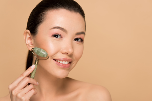 attractive naked asian woman making massage of face with jade roller isolated on beige - Photo, Image