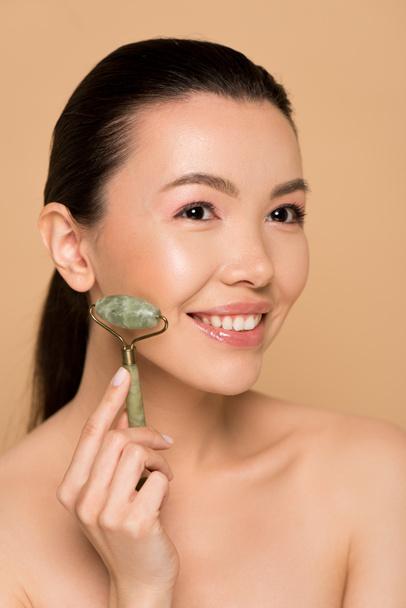 happy naked asian girl making massage of face with jade roller isolated on beige - Fotó, kép