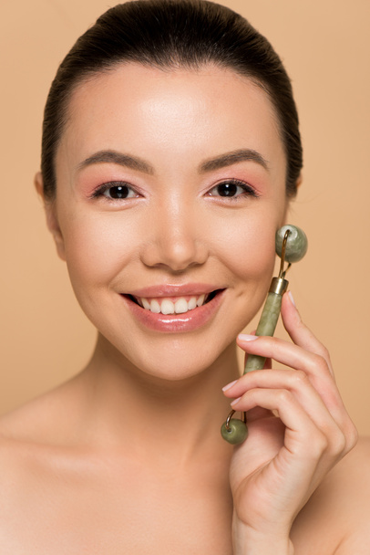 hermosa sonriente chica asiática desnuda haciendo masaje de cara con rodillo de jade aislado en beige
 - Foto, imagen
