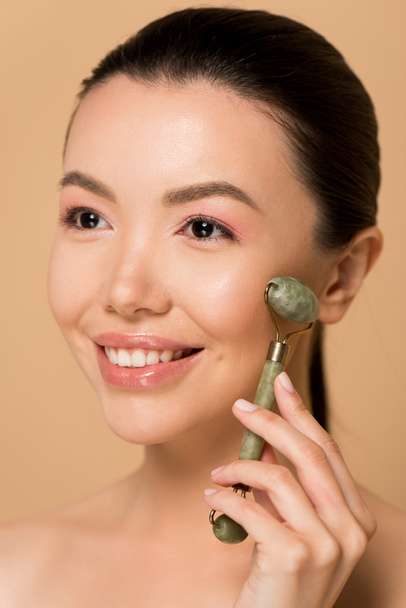 attractive happy naked asian girl making massage of face with jade roller isolated on beige - Photo, Image