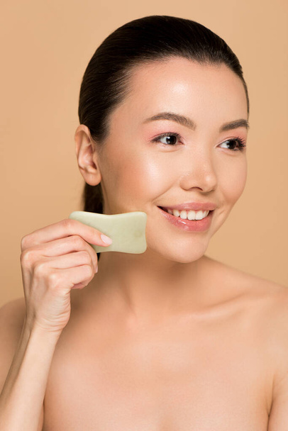 beautiful happy nude asian woman making massage of face with jade gua sha stone isolated on beige - Фото, изображение