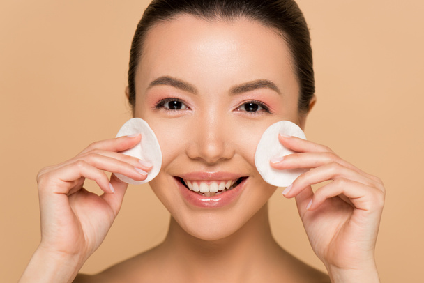 sonriente asiático mujer la eliminación de maquillaje de la cara con algodón almohadillas aislado en beige
 - Foto, Imagen