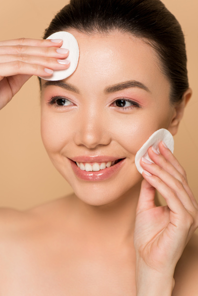 bela mulher asiática feliz removendo maquiagem do rosto com almofadas de algodão isolado no bege
 - Foto, Imagem