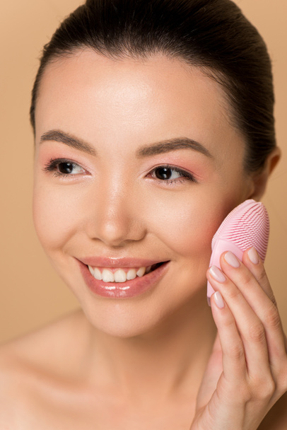 beautiful smiling naked asian girl using silicone cleansing facial brush isolated on beige - Photo, Image