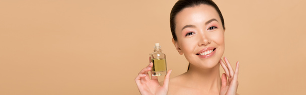 panoramisch schot van mooie vrolijke naakt aziatische meisje aanbrengen parfum op de huid geïsoleerd op beige - Foto, afbeelding