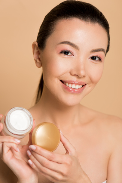 hermosa sonriente chica asiática desnuda sosteniendo contenedor de vidrio con crema cosmética aislado en beige
 - Foto, Imagen