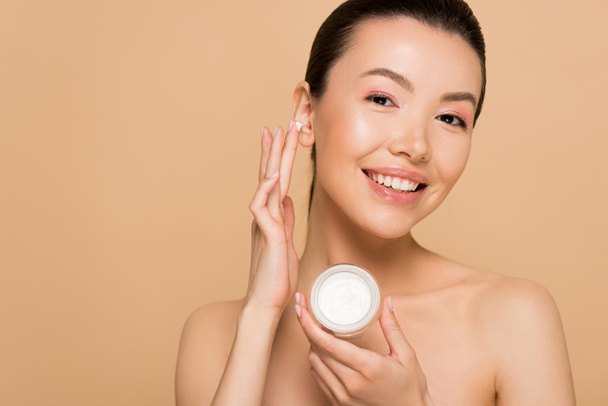 beautiful nude asian woman applying cosmetic cream on face isolated on beige  - Fotoğraf, Görsel