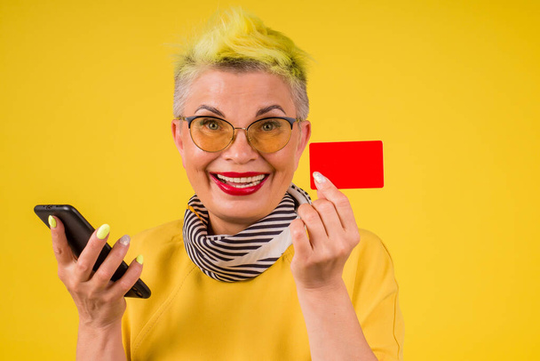 mature old woman with colored stylish yellow dyed hair and red lipstick lips in warm dress holding credit card and phone background studio:black friday online sale - Fotografie, Obrázek