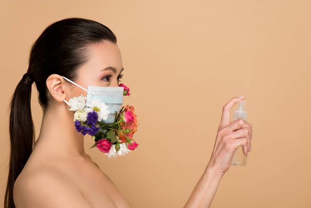 attractive nude asian girl in floral face mask holding antiseptic spray isolated on beige - Photo, Image