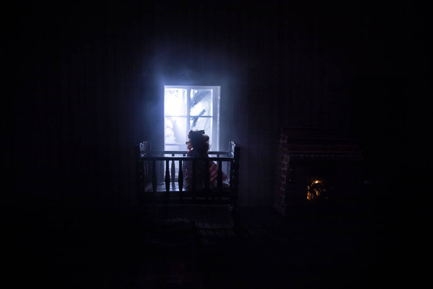 Old creepy eerie baby crib near window in dark room. Scary baby silhouette in dark. A realistic dollhouse living room with furniture and window at night. Selective focus - Photo, Image